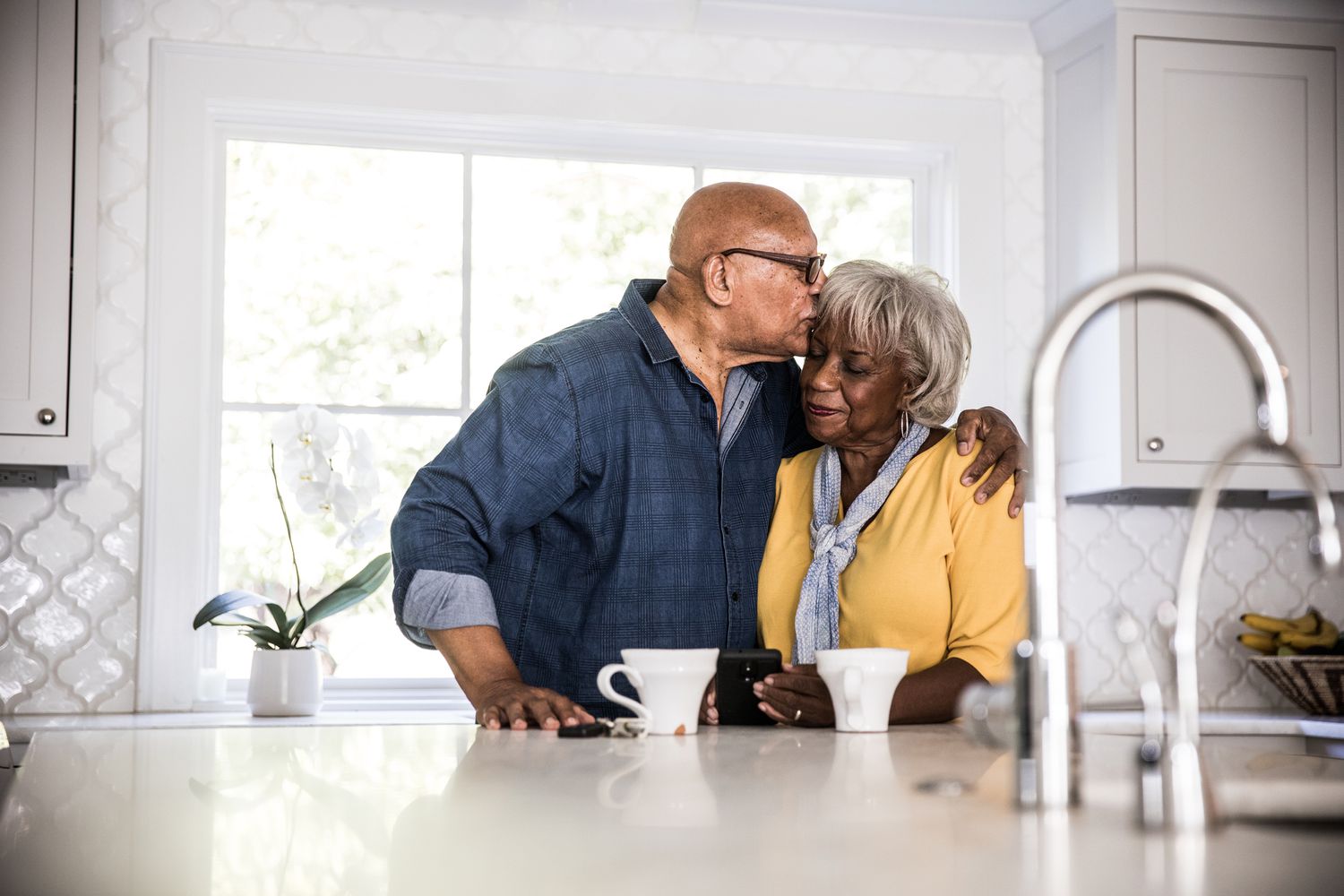 一个老男人吻了一个老女人的头，站在厨房里，柜台上放着咖啡杯