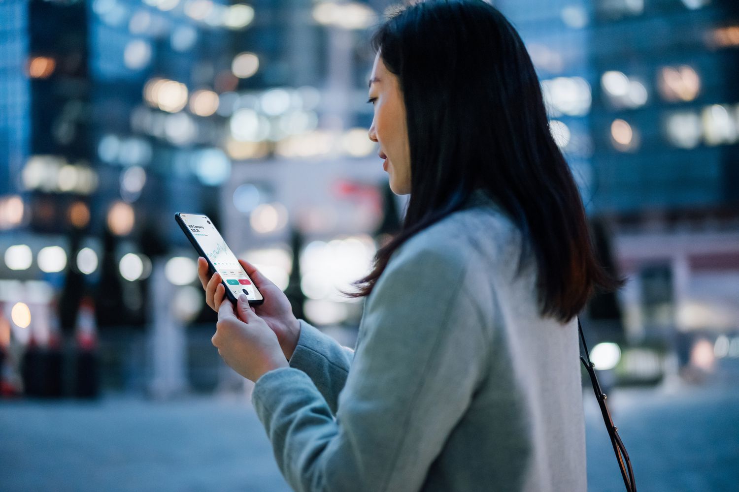 年轻的亚洲女商人在路上用智能手机上的移动应用程序检查金融股票市场分析，晚上在金融区的当代企业摩天大楼。