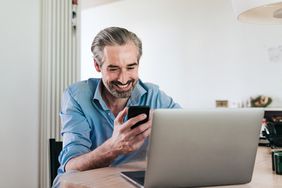 男人笑眯眯地看着手机,坐在笔记本电脑前