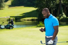 阳光球场上的高尔夫球手拿着球杆，看着智能手机