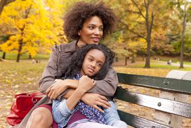一个母亲和女儿在秋天公园的长椅上坐着”width=