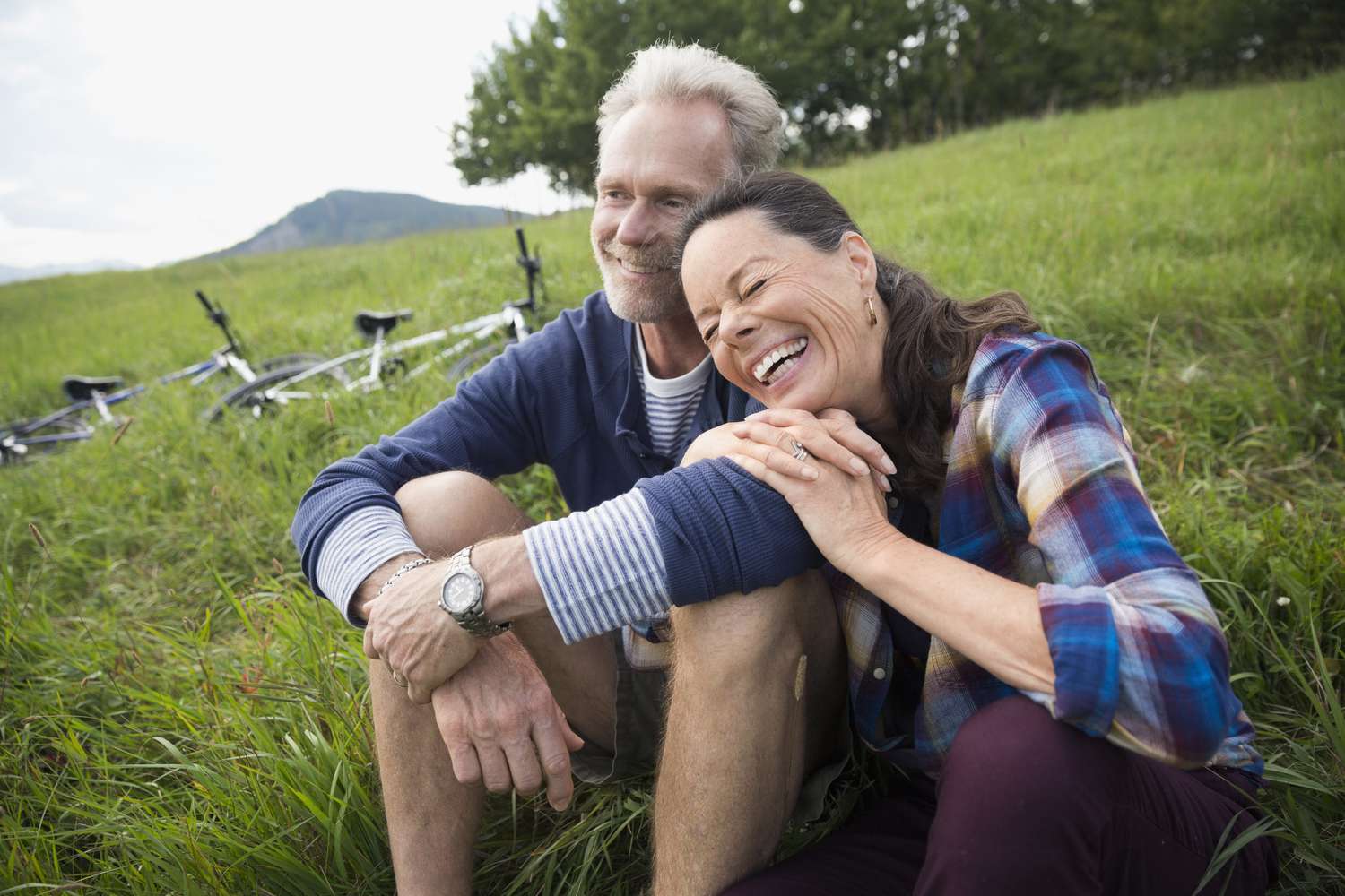 一对笑着的退休夫妇坐在草地上，旁边有两辆自行车。＂class=