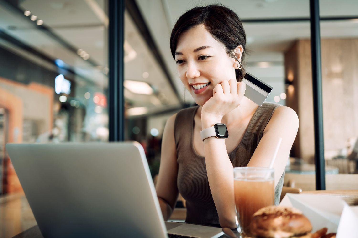 美丽微笑的年轻亚洲女子在咖啡馆吃饭，用笔记本电脑在网上购物，用信用卡在手进行移动支付＂class=