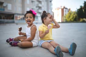 大热天，两个小女孩坐在人行道上，享受着融化的新奇冰淇淋。其中一人似乎正在经历“脑冻结”。