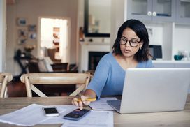 一名女子戴着眼镜，拿着笔记本电脑和计算器坐在厨房桌子旁