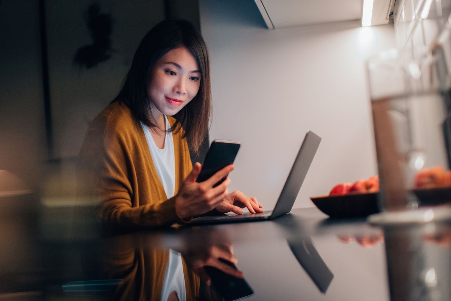 年轻的亚洲妇女看智能手机与笔记本电脑在厨房工作时在晚上在家”class=