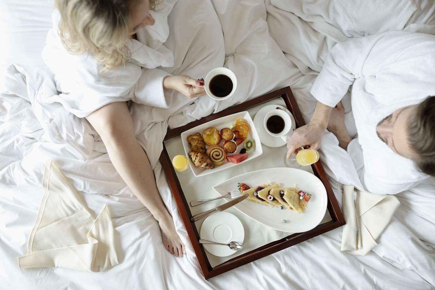 在这张俯拍的照片中，一男一女穿着长袍坐在酒店的床上，享受着有糕点、咖啡和果汁的欧式早餐。
