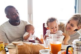 美丽年轻的跨种族家庭在家里，他们可爱的女儿和小儿子一起吃早餐。＂width=