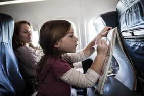 一个孩子坐在飞机上乘客的作品在一个彩色书,坐在她旁边的一个女人看起来。”>
          </noscript>
         </div>
        </div>
       </div>
       <div class=
