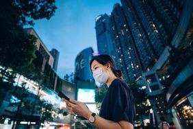 年轻的亚洲女子戴着防护口罩，晚上在市中心街道上使用智能手机，对面是霓虹灯商业招牌。日常生活的新常态。忙碌的生意