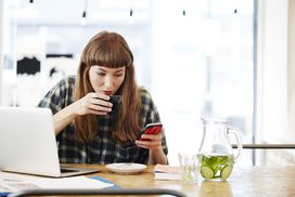 年轻女子检查她的手机而喝咖啡在她面前的笔记本电脑”width=