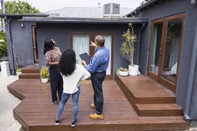 一位房地产经纪人向两位女士展示一套房子的特征。所有人都站在后院的露台上，背对着镜头看着屋顶。