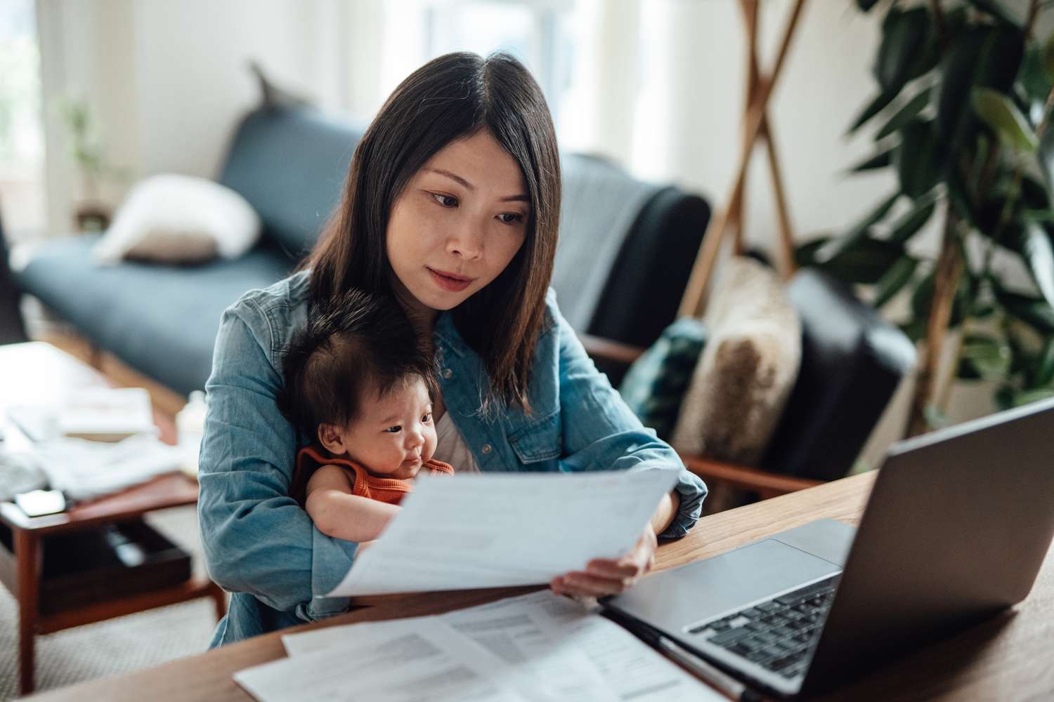 一名妇女抱着婴儿坐在办公桌前，面前是一台打开的笔记本电脑，她专注地看着手里的一份文件