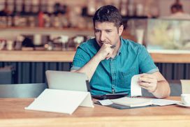 Concerned man at coffee shop looking at laptop with paperwork