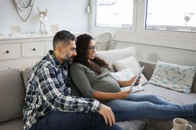 A man with a beard and a pregnant woman sit on a couch looking over their budget on a tablet