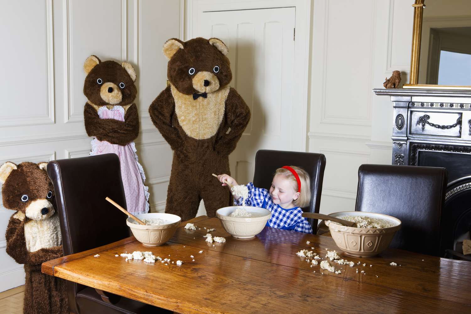 一个小女孩坐在被三只熊围着的桌子上，用碗吃东西