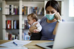 图为，带着婴儿、戴着口罩的一名妇女在家中盯着笔记本电脑屏幕。
