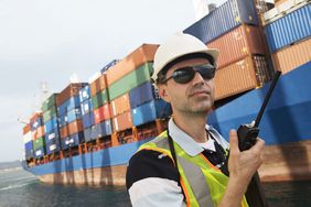 一名戴着安全帽的工人手持通信设备，背景是一艘货船。