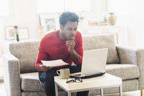 A man sitting on a couch leans forward and reviews documents, an open laptop on the coffee table in front of him.