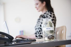 一个女人在桌子前工作，桌上放着一罐标有401k的现金。＂width=