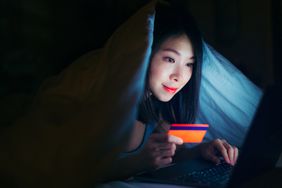 特写:年轻的亚洲女性用信用卡和笔记本电脑在网上购物，盖上羽绒被，躺在黑暗的床上。