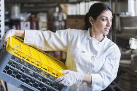 一个女人在一个商业厨房工作。她穿着一件白色制服,乳胶手套,拿着一个塑料箱