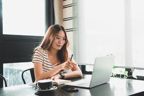 Woman sits at a table with a coffee cup looking at a laptop