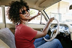 一位留着自然发型、戴着墨镜的微笑女子开着她的车