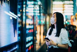 戴着防护口罩的女商人在证券交易所的显示屏前查看智能手机上的金融交易数据＂width=