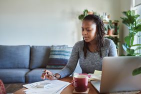 一个女人坐在家里的桌子旁，用一堆纸和一台笔记本电脑计算税收