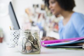 一个女人在桌子上的笔记本电脑上工作，她有一小罐零钱作为应急基金。
