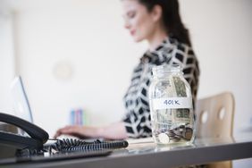 一个女人在桌子前工作，桌上放着一罐标有401k的现金。＂>
          </noscript>
         </div>
        </div>
       </div>
       <div class=