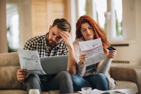 Worried couple seated on a couch looking over their paper bills; laptop on manâs lap