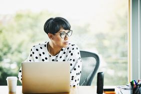一位戴着眼镜、穿着波点衬衫的女士坐在笔记本电脑前，考虑着保险的可携带性，目光转向了一边