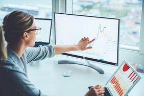 一位女士指着两个不同的电脑屏幕，上面有彩色的股票图表。