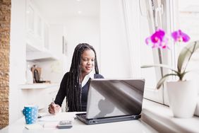 一个留着脏辫的女人坐在厨房桌子旁，用笔记本电脑计算所得税