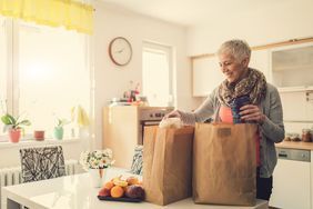 一名妇女微笑着站在厨房桌子上装满杂货的购物纸袋旁