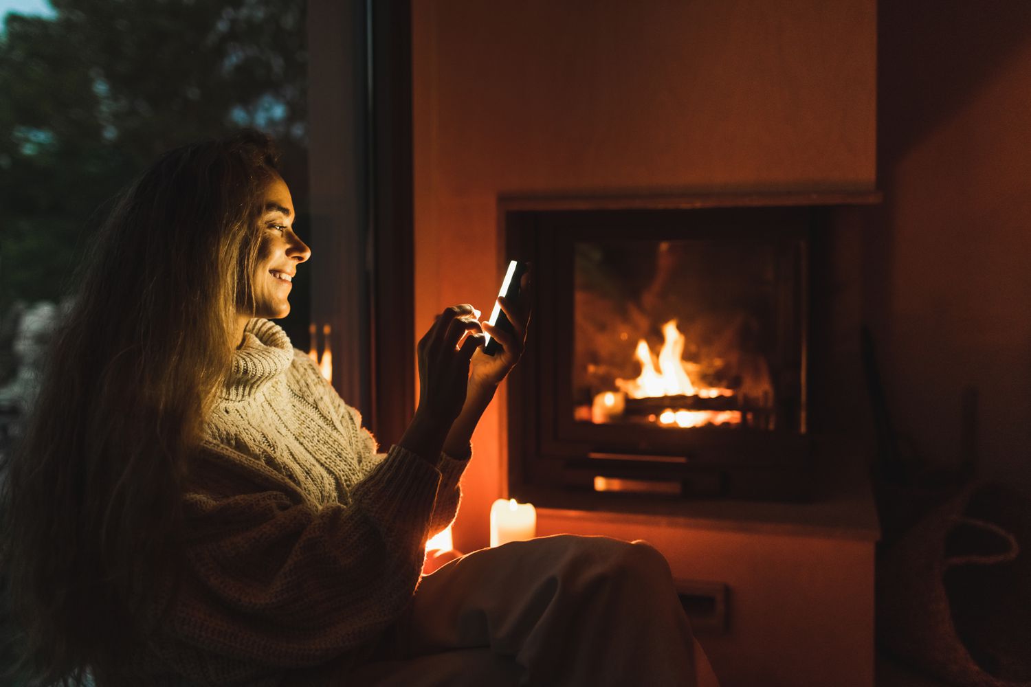 微笑的女人坐在窗台上的手机靠近壁炉在晚上
