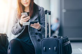 一名女子拿着护照，坐在机场的黑色滚动行李箱旁，看着自己的手机和登机牌。