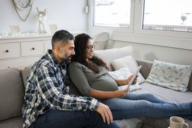 一个留胡子的男人和一个怀孕的女人坐在沙发上，用平板电脑查看他们的预算”width=