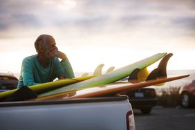 一个婴儿潮时期出生的人在冲浪了一整天后，靠在一辆皮卡车后面的一堆冲浪板上，想知道他是否应该多存钱，少冲浪。