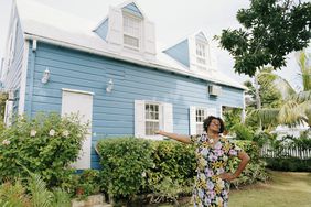 一位穿着碎花裙的妇女自豪地指着她那有白色百叶窗和可爱花园的亮蓝色房子