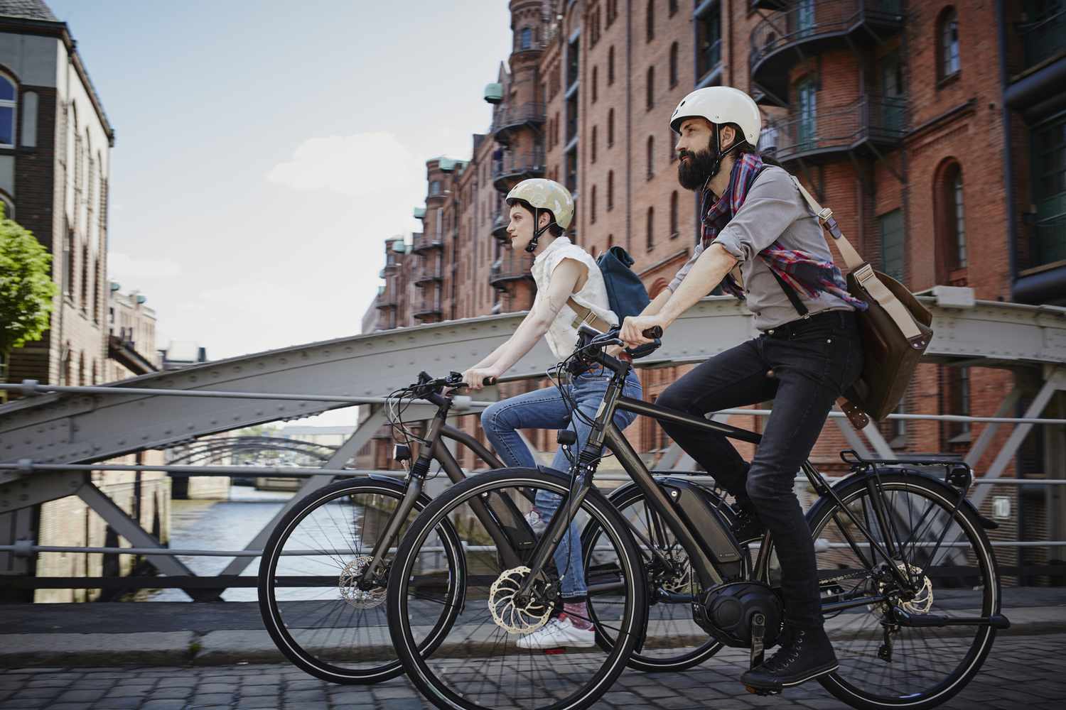 一对夫妇戴着头盔，背着背包，骑着电动自行车走过一座桥，背景是砖墙建筑
