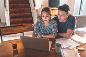 A young couple sitting together at a kitchen table together in front of a laptop looking intently at the screen