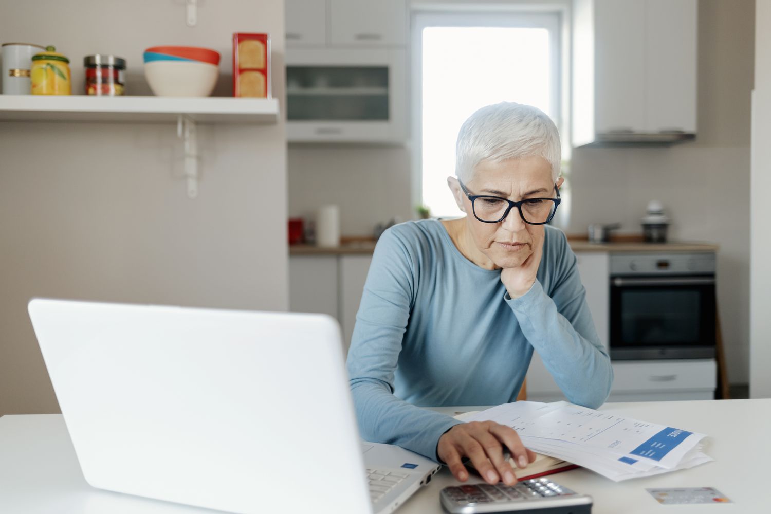 一名老年妇女在厨房桌子上用计算器工作，笔记本电脑和文件就在旁边