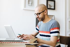 戴眼镜的秃头男子坐在桌前，手里拿着笔记本电脑和笔