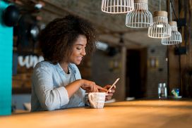 一个人在咖啡馆边喝咖啡边用手机看社交媒体＂width=