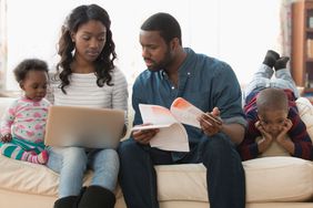 Stressed parents paying bills on laptop while two young children try to get their attention.