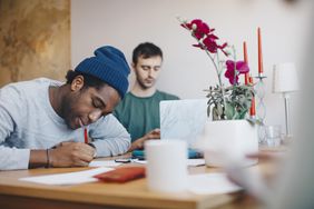 young man in blue beanie studying at table, while another young man in green shirt studies behind him