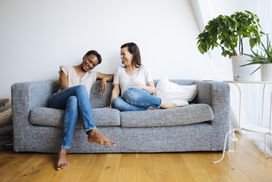 两个快乐放松的女人坐在家里的沙发上＂width=